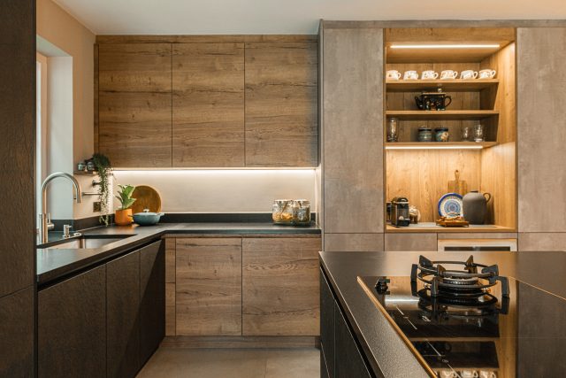Contemporary kitchen with wood-effect cabinetry, black worktops, LED under-cabinet lighting and open display shelving, Ashley Jay Kitchens
