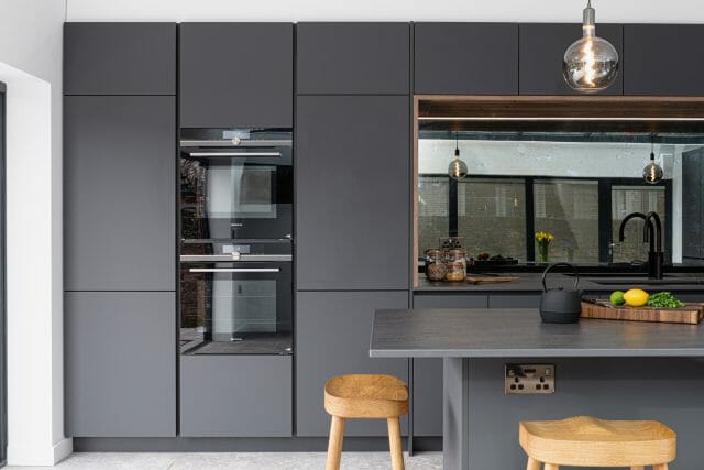 Contemporary grey Ashley Jay kitchen with handleless cabinetry, integrated ovens, island and mirrored splashback
