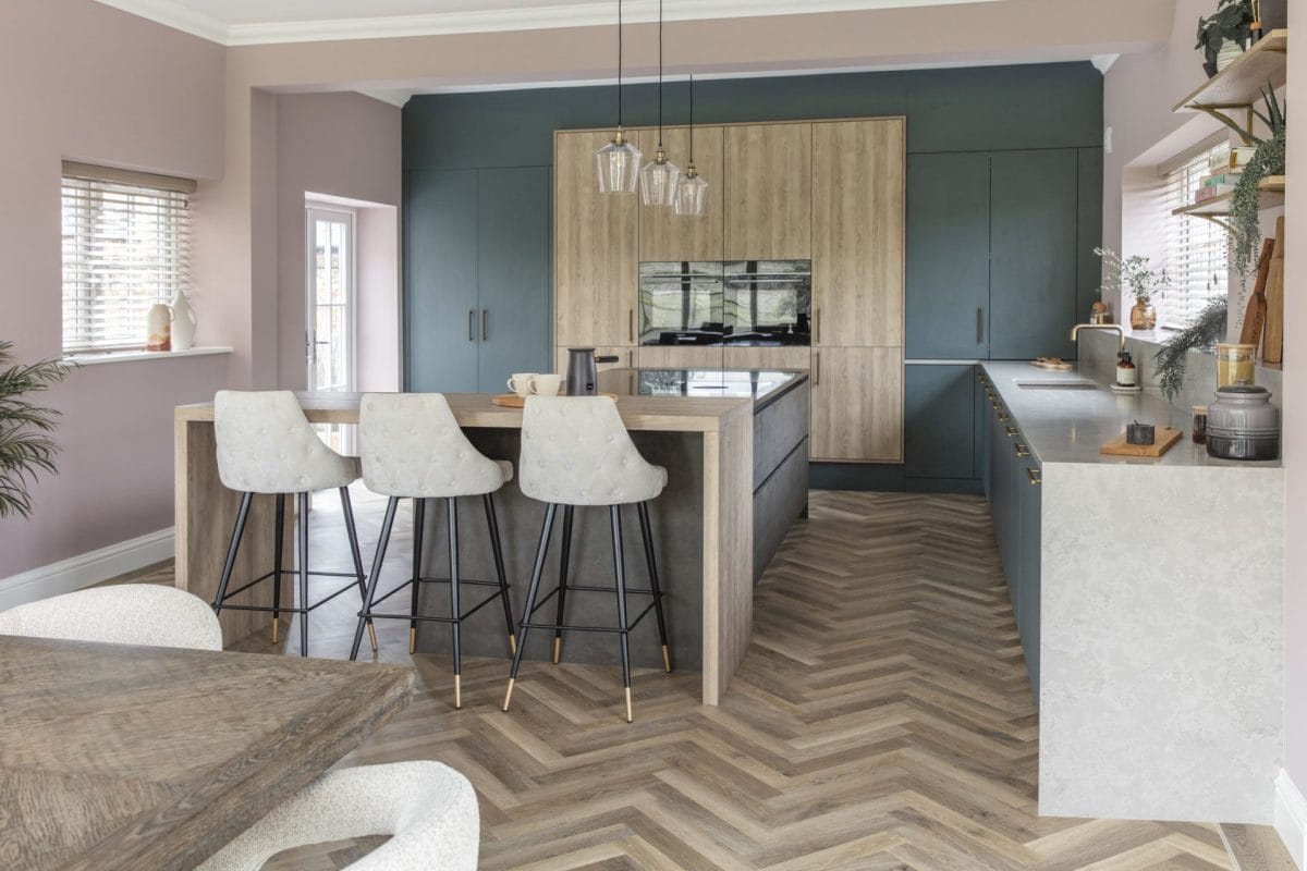 Modern kitchen featuring deep green cabinetry, natural wood finishes and a spacious peninsula with breakfast bar seating, complemented by warm neutral tones and herringbone flooring.